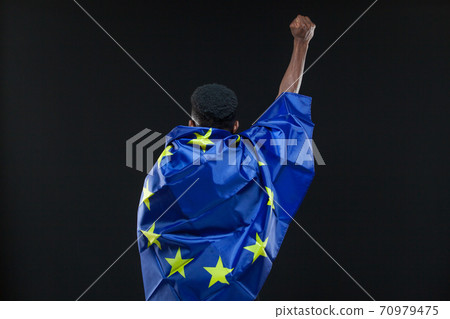 Back view of young african american man wrapped in the flag of the European Union standing with raised fist isolated on black background. Concept of the European Union, unification, integration Back view of young african american man wrapped in the flag of the European Union standing with raised fist isolated on black background. Concept of the European Union, unification, integration 70979475