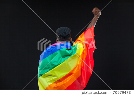 Back view of young african american man wrapped in a rainbow flag standing with raised fist isolated on black background. Concept of The LGBT community, minority rights, protection of human rights Back view of young african american man wrapped in a rainbow flag standing with raised fist isolated on black background. Concept of The LGBT community, minority rights, protection of human rights 70979478