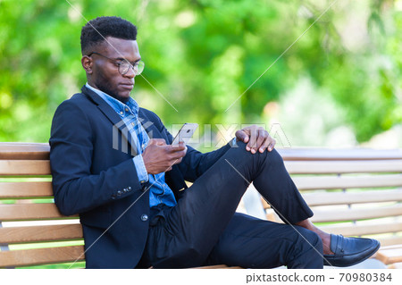 Young African entrepreneur is focused on work issues and communicating on a mobile phone for business deals sitting on a bench in a summer park 70980384