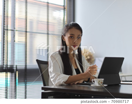 Female office worker looking into camera while working with tablet in office room Female office worker looking into camera while working with tablet in office room 70980717