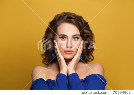 Elegant woman with blue eyeshadow makeup wearing blue top on yellow background 70994902