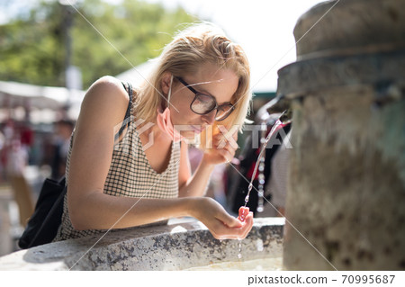 Thirsty young casual cucasian woman wearing medical face mask drinking water from public city fountain on a hot summer day. New social norms during covid epidemic Thirsty young casual cucasian woman wearing medical face mask drinking water from public city fountain on a hot summer day. New social norms during covid epidemic 70995687