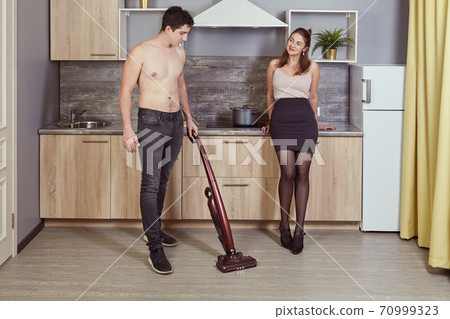White shirtless man cleans room with wireless vacuum cleaner with young woman near. 70999323