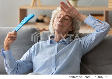 Tired senior woman waving fan feel hot sit on sofa Tired senior woman waving fan feel hot sit on sofa 71007968