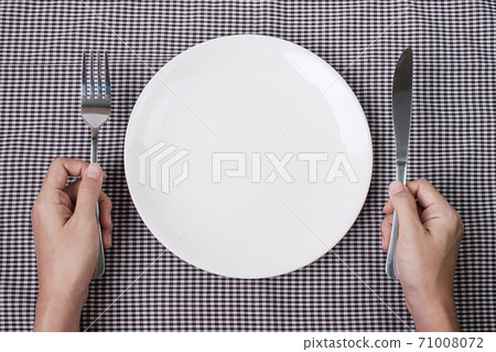 Hands holding knife and fork above white plate on table background., dieting, weight loss, dining and kitchenware concept Hands holding knife and fork above white plate on table background., dieting, weight loss, dining and kitchenware concept 71008072
