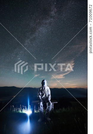 Illuminated man wearing white space suit and helmet sitting on a bench on a hill at night 71009672