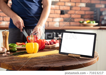 Tablet on the kitchen table and man preparing delicious and healthy food on background. Tablet screen with empty space. Tablet on the kitchen table and man preparing delicious and healthy food on background. Tablet screen with empty space. 71013217