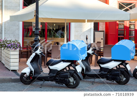 A moped for delivering food from the restaurant to the house is parked at the front of the cafe with an umbrella and tables decorated with flowerpots with flowers. A moped for delivering food from the restaurant to the house is parked at the front of the cafe with an umbrella and tables decorated with flowerpots with flowers. 71018939