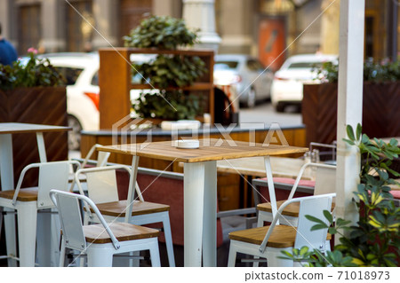 Empty table in the summer terrace cafe, a wooden tabletop and an iron leg against the background of chairs and shelves with plants. Empty table in the summer terrace cafe, a wooden tabletop and an iron leg against the background of chairs and shelves with plants. 71018973