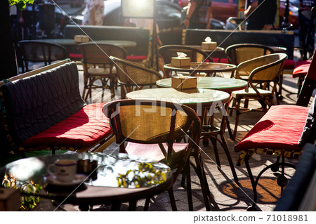 Outdoor cafe with soft seats and glass tables with a sun flare, an empty cafe and people with cars in the background of the restaurant. 71018981