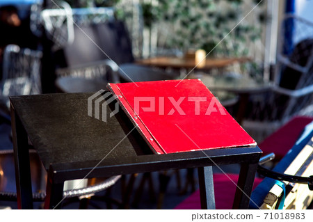 A black wooden stand on which lies a red folder with a restaurant menu on the background of empty tables nobody. 71018983