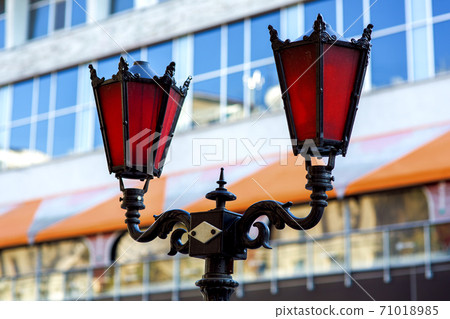 Street lamp lighting forged from metal in black and red glass, a lantern against the background of the facade of the building close-up. Street lamp lighting forged from metal in black and red glass, a lantern against the background of the facade of the building close-up. 71018985