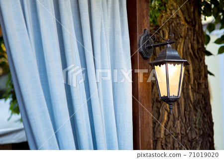 a street lamp on a brown pillar against the background of blue curtains and a tree trunk, an antique semi-antique decorative lantern with a warm light. a street lamp on a brown pillar against the background of blue curtains and a tree trunk, an antique semi-antique decorative lantern with a warm light. 71019020
