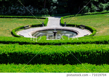 a stone round fountain with water surrounded by a walkway of tiles with wooden benches and a hedge of evergreen boxwood bushes on a sunny summer day. a stone round fountain with water surrounded by a walkway of tiles with wooden benches and a hedge of evergreen boxwood bushes on a sunny summer day. 71019704