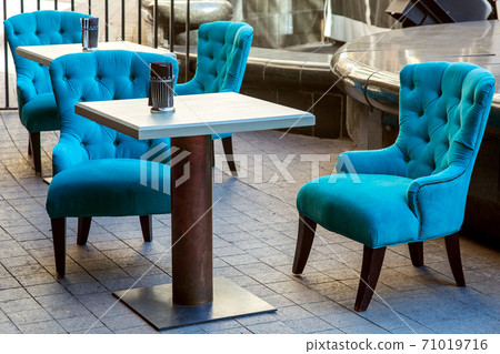 an outdoor cafe with square wooden tables and an iron leg and upholstered armchairs with high backs and sheathed with fabric installed on the sidewalk of stone tiles. an outdoor cafe with square wooden tables and an iron leg and upholstered armchairs with high backs and sheathed with fabric installed on the sidewalk of stone tiles. 71019716