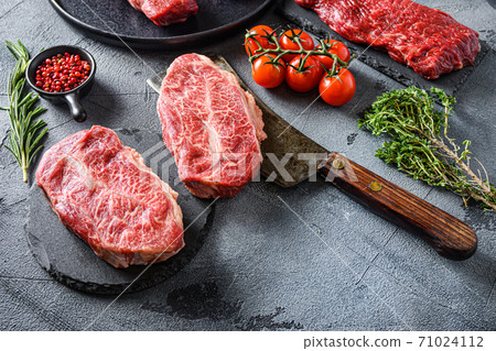 Organic feather steak steak on chopping board, marbled beef over other alternative cuts with herbs tomatoes peppercorns over grey stone surface background side view close up selective focus new wide 71024112