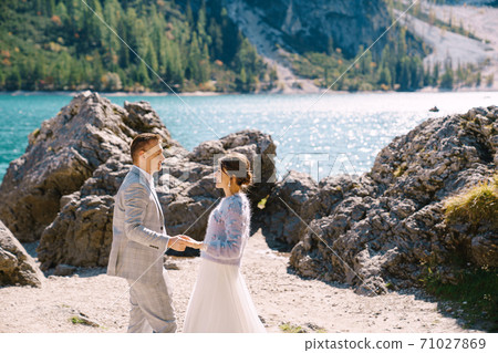 Bride and groom stand against the backdrop of stones overlooking Lago di Braies in Italy. Destination wedding in Europe, on Braies lake. Loving newlyweds walk against the backdrop of amazing nature. Bride and groom stand against the backdrop of stones overlooking Lago di Braies in Italy. Destination wedding in Europe, on Braies lake. Loving newlyweds walk against the backdrop of amazing nature. 71027869