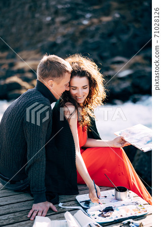 A guy hugs a girl artist who paints a picture in the album with watercolors. Near Ehsaraurfoss waterfall, Ehsarau River, Thingvedlir National Park in Iceland. A guy hugs a girl artist who paints a picture in the album with watercolors. Near Ehsaraurfoss waterfall, Ehsarau River, Thingvedlir National Park in Iceland. 71028126