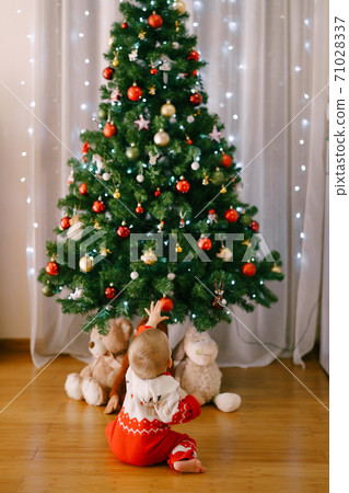 Baby in a funny knitted costume is reaching for a Christmas ornament on a Christmas tree 71028337