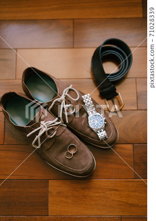 Brown mens suede shoes with wristwatch, belt and wedding rings of the bride and groom on a wooden texture. Brown mens suede shoes with wristwatch, belt and wedding rings of the bride and groom on a wooden texture. 71028339