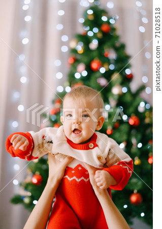 Toddler in a red-and-white Christmas bodysuit in her mother's arms in front of a Christmas tree 71028518