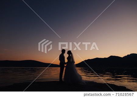the bride and the groom standing on the pier near water and holding hands at the sunset 71028567