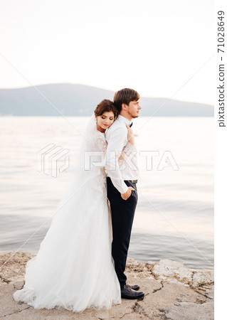 the bride hugs the groom, resting her head on his back on the pier near water the bride hugs the groom, resting her head on his back on the pier near water 71028649