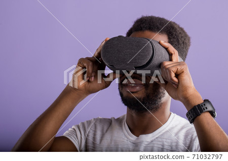 Handsome young African man in VR headset standing against purple background Handsome young African man in VR headset standing against purple background 71032797