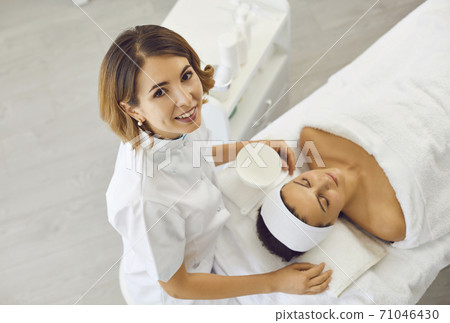Cosmetologist or dermatologist looking up at camera near realxing woman in beauty salon Cosmetologist or dermatologist looking up at camera near realxing woman in beauty salon 71046430
