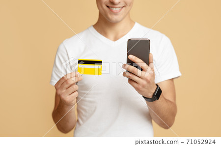 Cheerful millennial man in white t-shirt looks at smartphone and holds credit card, pays for purchase 71052924