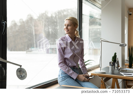 Dreamy young businesswoman sitting on windowsill in office. 71053373