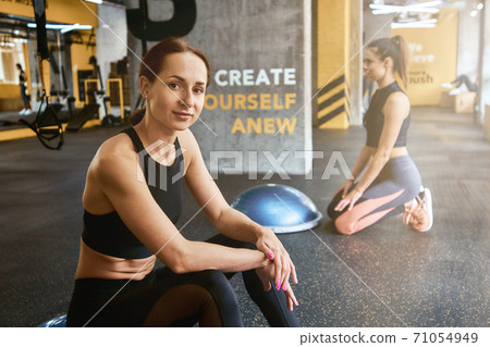 Young beautiful woman in sportswear smiling at camera while working out with friend at gym, using special fitness equipment Young beautiful woman in sportswear smiling at camera while working out with friend at gym, using special fitness equipment 71054949