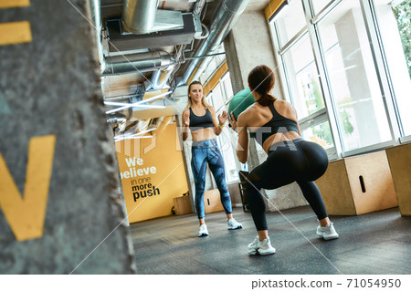 Rear view of a young athletic woman in sportswear squatting with weight ball at gym, working out with personal trainer 71054950