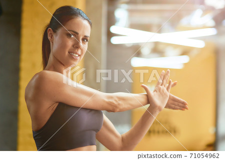 Young beautiful fitness woman in sportswear stretching muscles and looking at camera, warming up before workout at gym 71054962