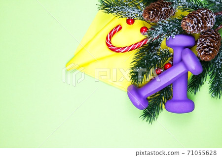 Christmas fitness. Healthy and active lifestyles greeting card concept. Purple dumbbells, yellow rubber band, candy and fir-tree on pink 71055628