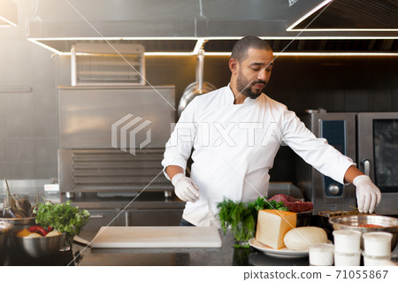 Handsome young African chef standing in professional kitchen in restaurant preparing a meal of meat and cheese vegetables. 71055867