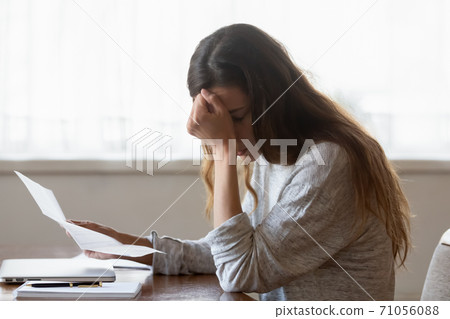 Upset woman sitting at desk holding letter with bad news Upset woman sitting at desk holding letter with bad news 71056088