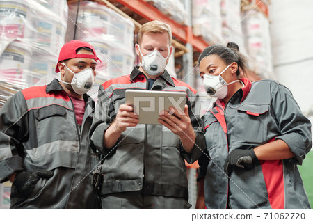 Group of warehouse workers in respirators scrolling through assortment of goods 71062720