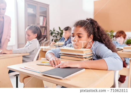 Bored mixed-race schoolgirl in casualwear keeping chin on top of stack of books Bored mixed-race schoolgirl in casualwear keeping chin on top of stack of books 71062811