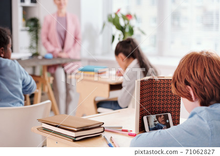 Contemporary pupil with smartphone sitting by desk and pretending to read book 71062827