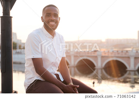 Happy young African businessman in white shirt and brown pants sitting by river Happy young African businessman in white shirt and brown pants sitting by river 71063142