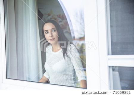 Woman staying indoors behind the window of her house, looking outside. Woman staying indoors behind the window of her house, looking outside. 71069891