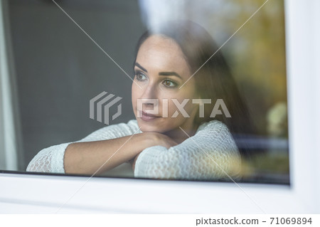 Depressed woman looks out from behind the window with a sad facial expression Depressed woman looks out from behind the window with a sad facial expression 71069894