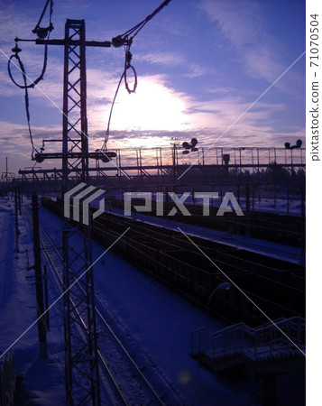 Winter northern short day. Low polar sun. Railway in the snow. Trains, freight cars and containers are on the rails of railway tracks. Silhouette of a high voltage power line. 71070504
