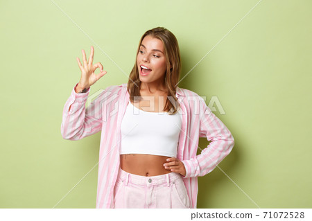 Portrait of attractive stylish woman with blond short hairstyle, wearing pink shirt with jeans, showing okay sign in approval, recommend something good, standing over green background 71072528
