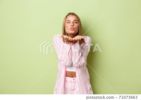 Portrait of beautiful romantic woman with blond short hair, blowing air kiss at camera express sympathy and love, standing over green background 71073663