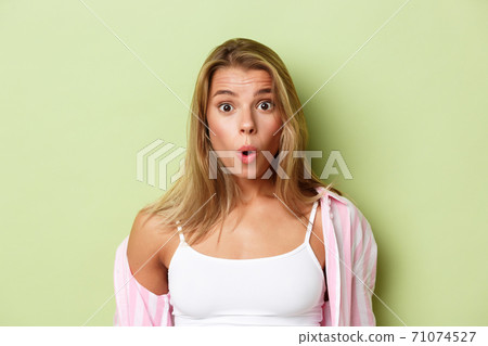 Close-up of attractive blond woman in pink shirt, looking surprised, saying wow fascinated, standing over green background 71074527