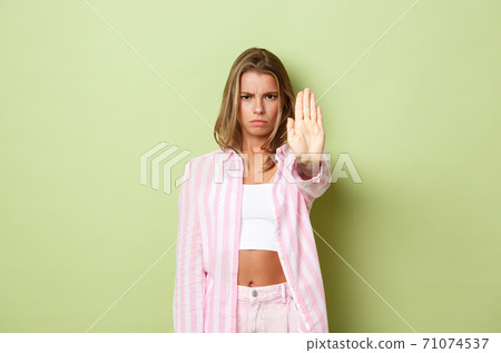 Portrait of serious and disappointed blond female model, showing stop sign and frowning, disapprove action, forbid something bad, standing over green background and saying no 71074537