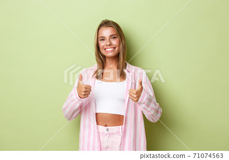 Portrait of beautiful woman with perfect white smile, showing her support and approval, make thumbs-up, standing over green background Portrait of beautiful woman with perfect white smile, showing her support and approval, make thumbs-up, standing over green background 71074563