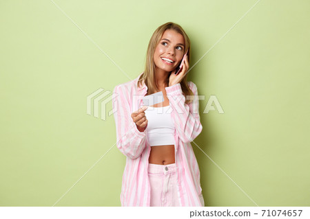 Image of young blond woman making a phone call and holding credit card, order something, standing over green background 71074657